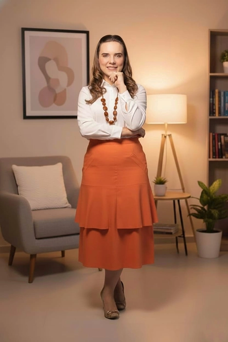 foto da Elizângela em pé, sorrindo, com o rosto apoiado na mão, vestindo blusa branca e saia bege