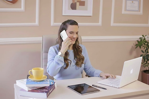 foto da elizângela em sentada, atrás de uma mesa com um notebook, falando no celular, com um livro à frente e outros 3 ao lado, empilhados, vestindo blusa azul, sorridente